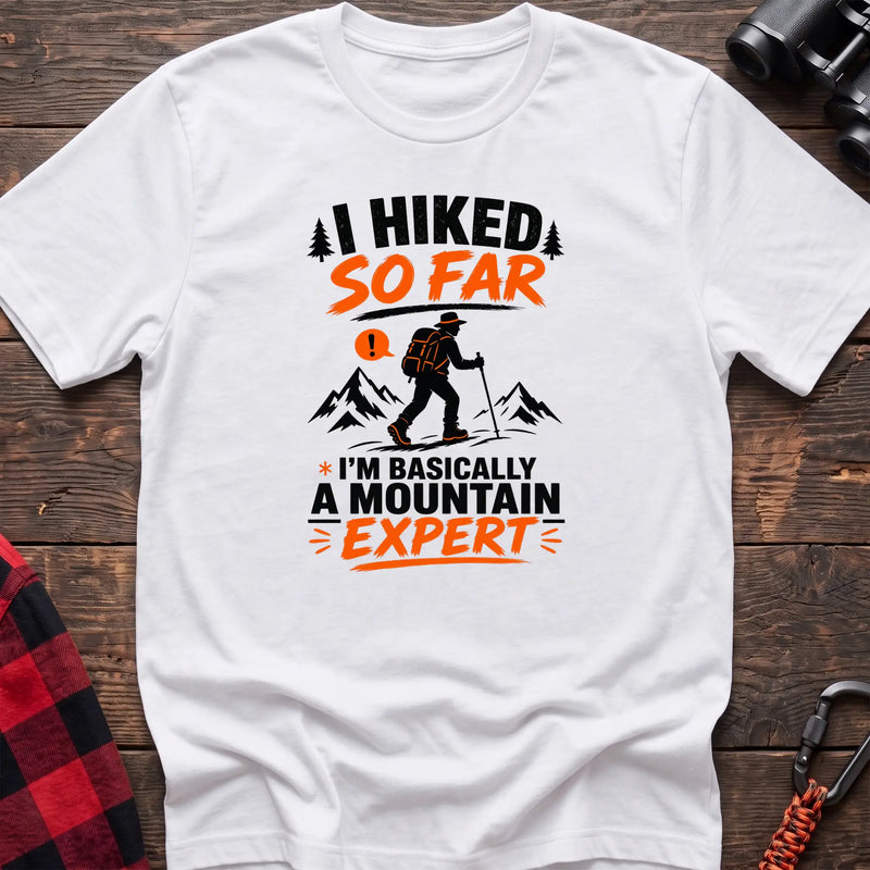White cotton t-shirt with I Hiked So Far I'm Basically a Mountain Expert graphic of a hiker with backpack and trekking pole, flat lay on rustic wood with hiking gear