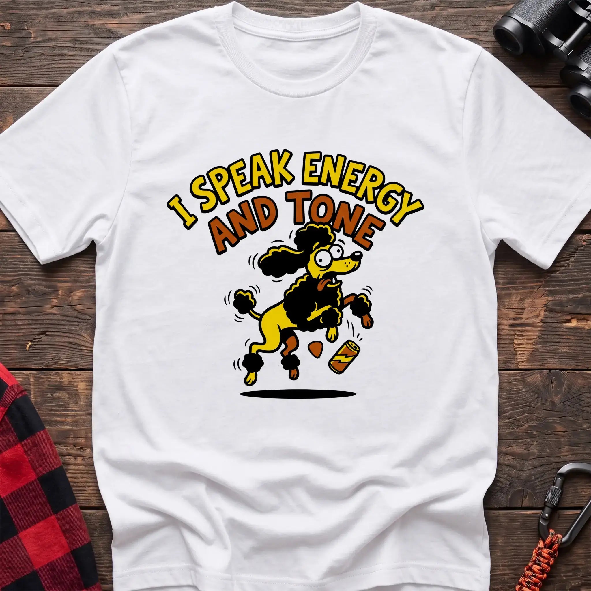 White t-shirt with a cartoon yellow and black poodle jumping excitedly next to a falling energy drink can, with the arched text “I SPEAK ENERGY AND TONE,” laid flat on a wooden surface.
