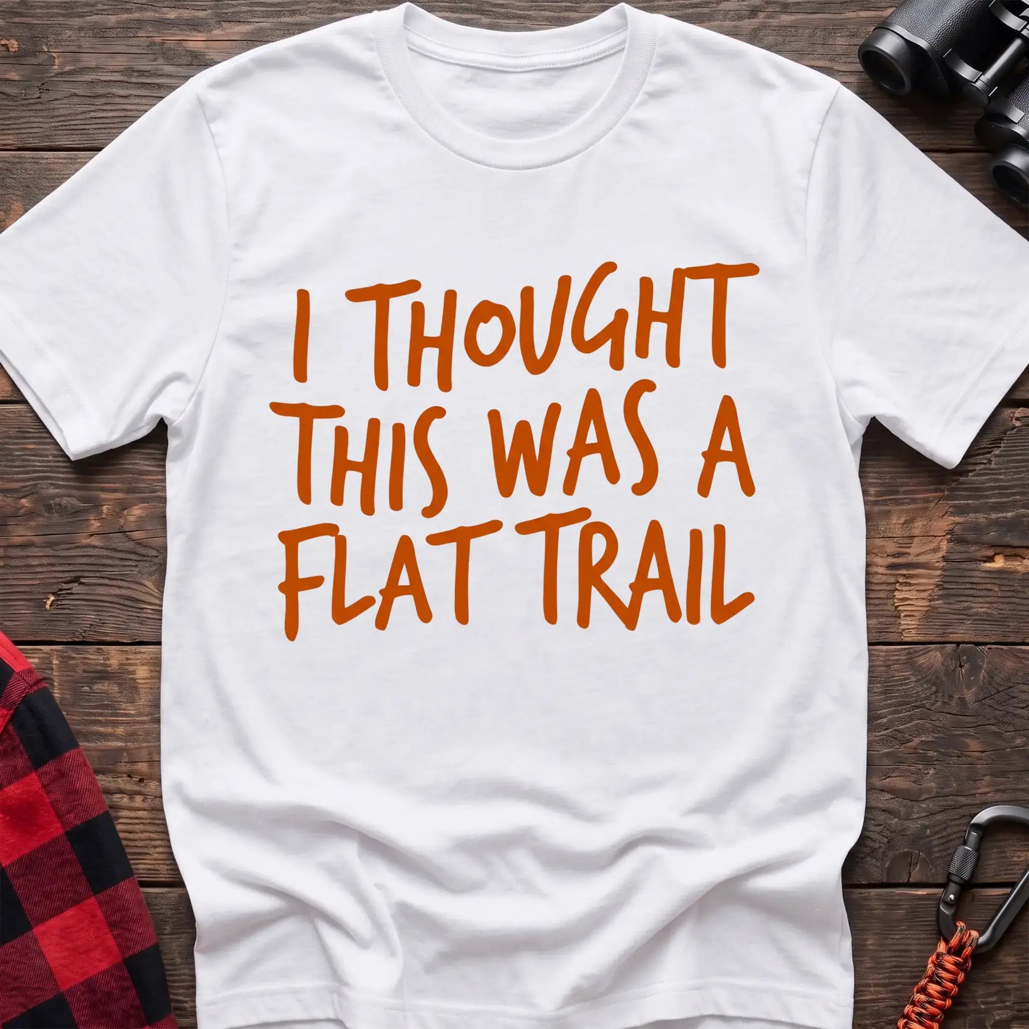 White t-shirt with bold orange handwritten-style text reading “I THOUGHT THIS WAS A FLAT TRAIL,” laid flat on a wooden surface.