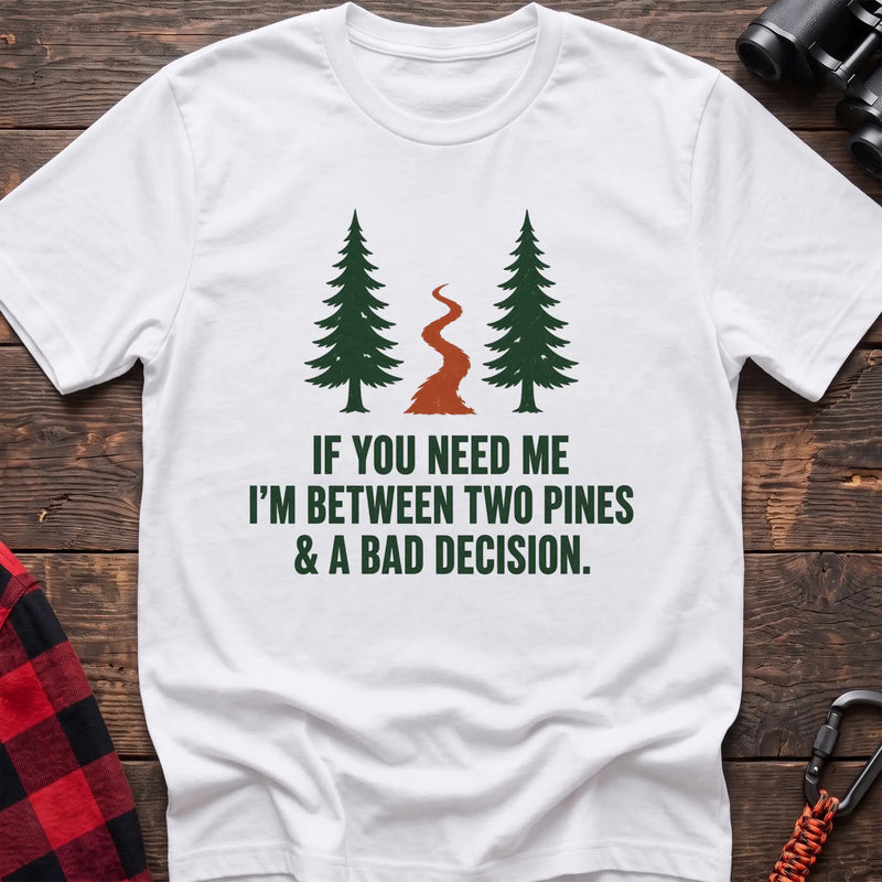 White cotton t-shirt with two pines and a trail graphic and If You Need Me I’m Between Two Pines and a Bad Decision text on rustic wood