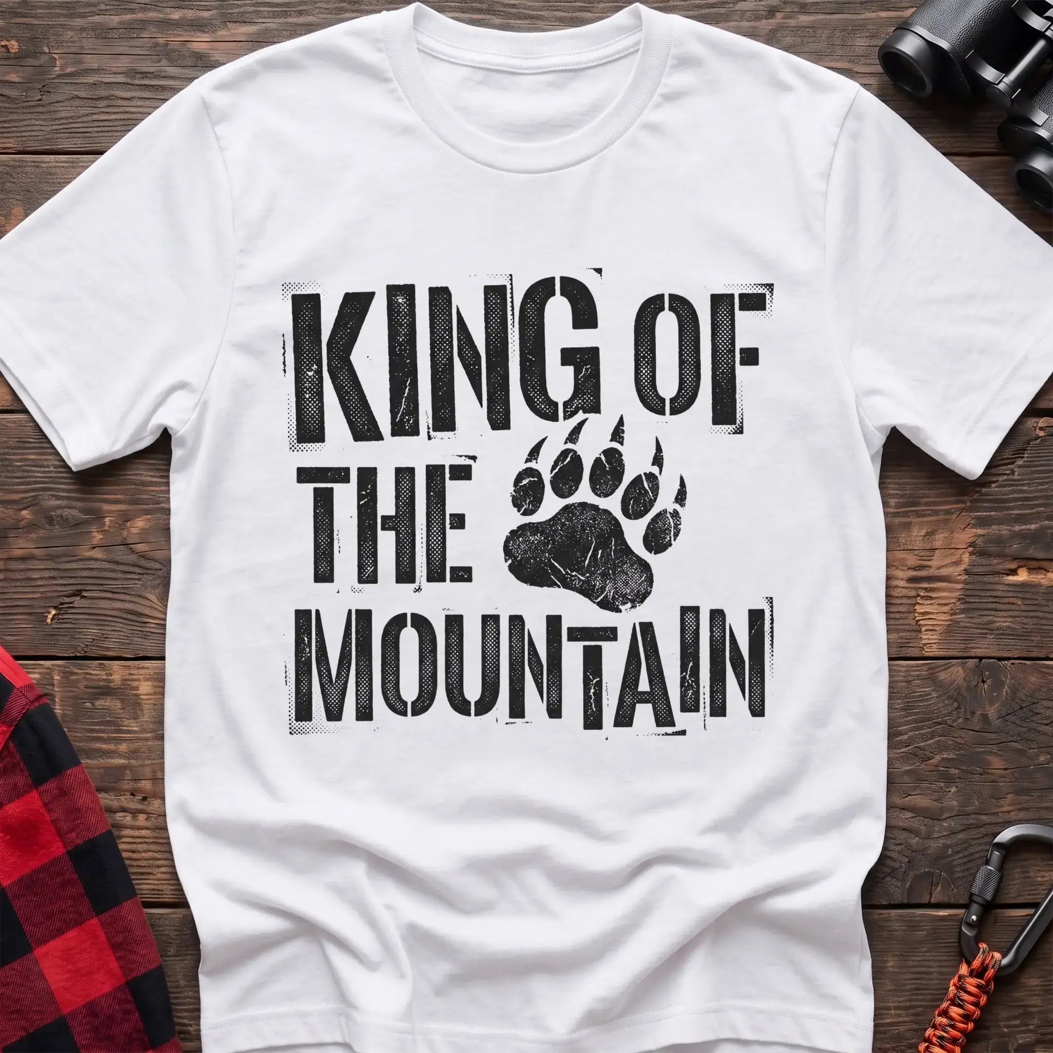 White t-shirt with distressed black text reading “KING OF THE MOUNTAIN” and a large bear paw print integrated into the design, laid flat on a wooden surface.