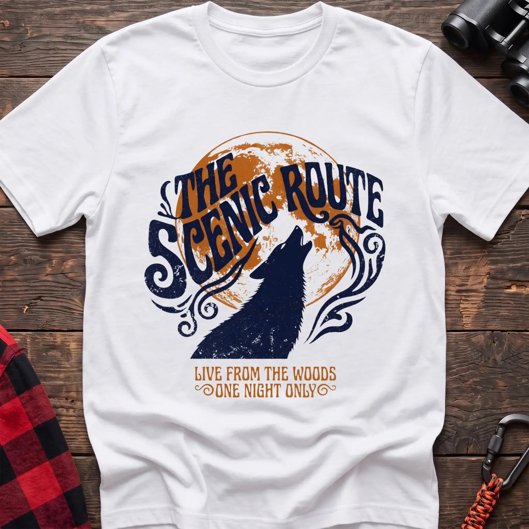White t-shirt with a dark blue howling wolf silhouette against an orange circle, with the arched text “THE SCENIC ROUTE” above and “LIVE FROM THE WOODS / ONE NIGHT ONLY” below, laid flat on a wooden surface.