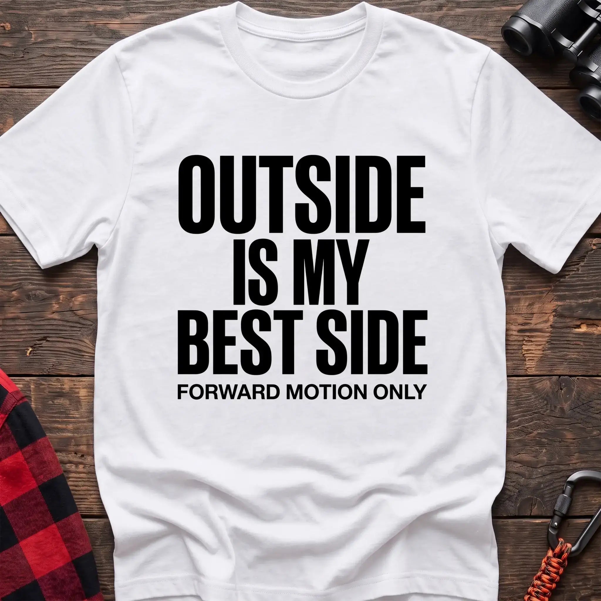 White t-shirt with bold black text reading “OUTSIDE IS MY BEST SIDE” and “FORWARD MOTION ONLY” below it, laid flat on a wooden surface.