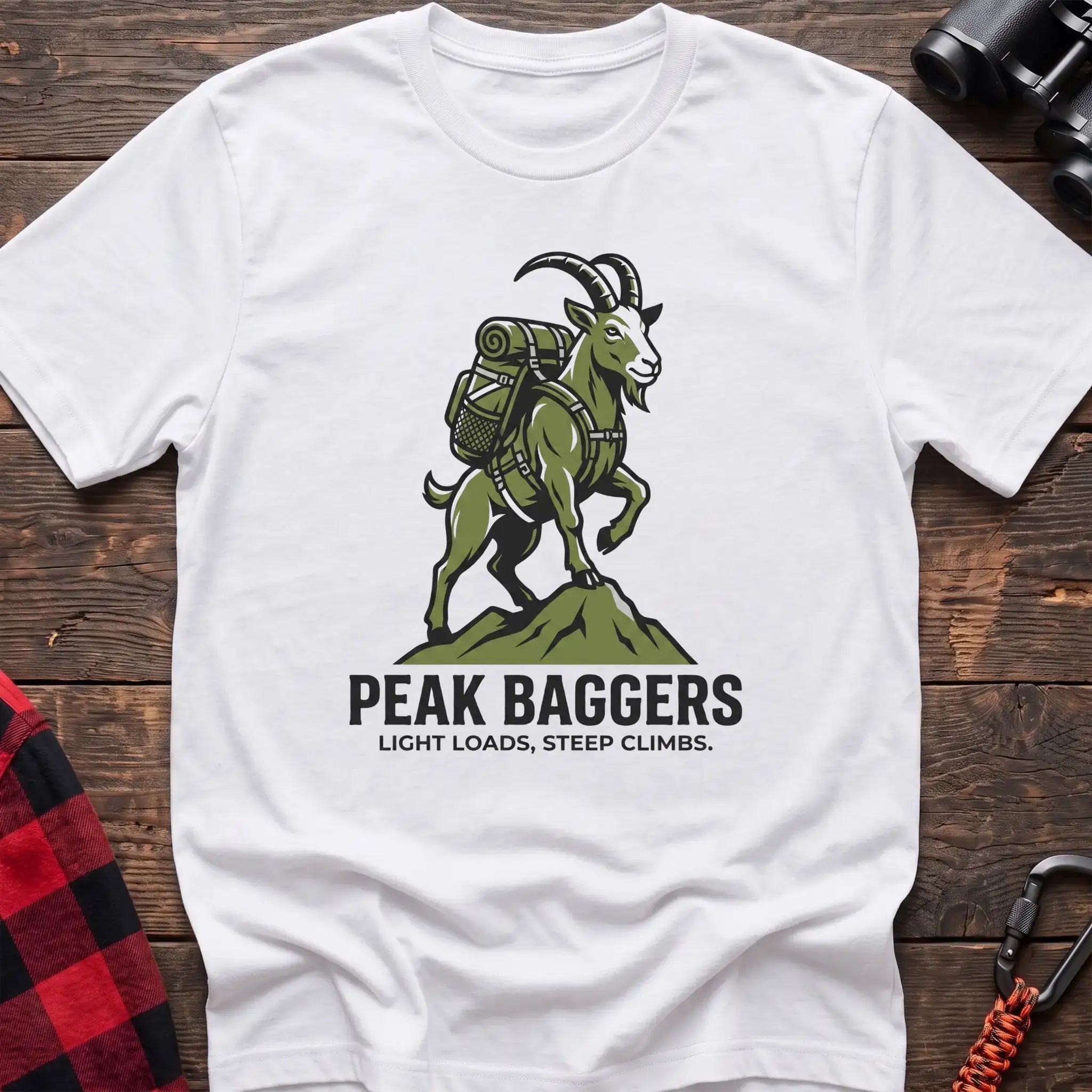 White t-shirt with an illustration of a mountain goat wearing a backpack standing on a peak, with the text “PEAK BAGGERS” and “LIGHT LOADS, STEEP CLIMBS,” laid flat on a wooden surface.