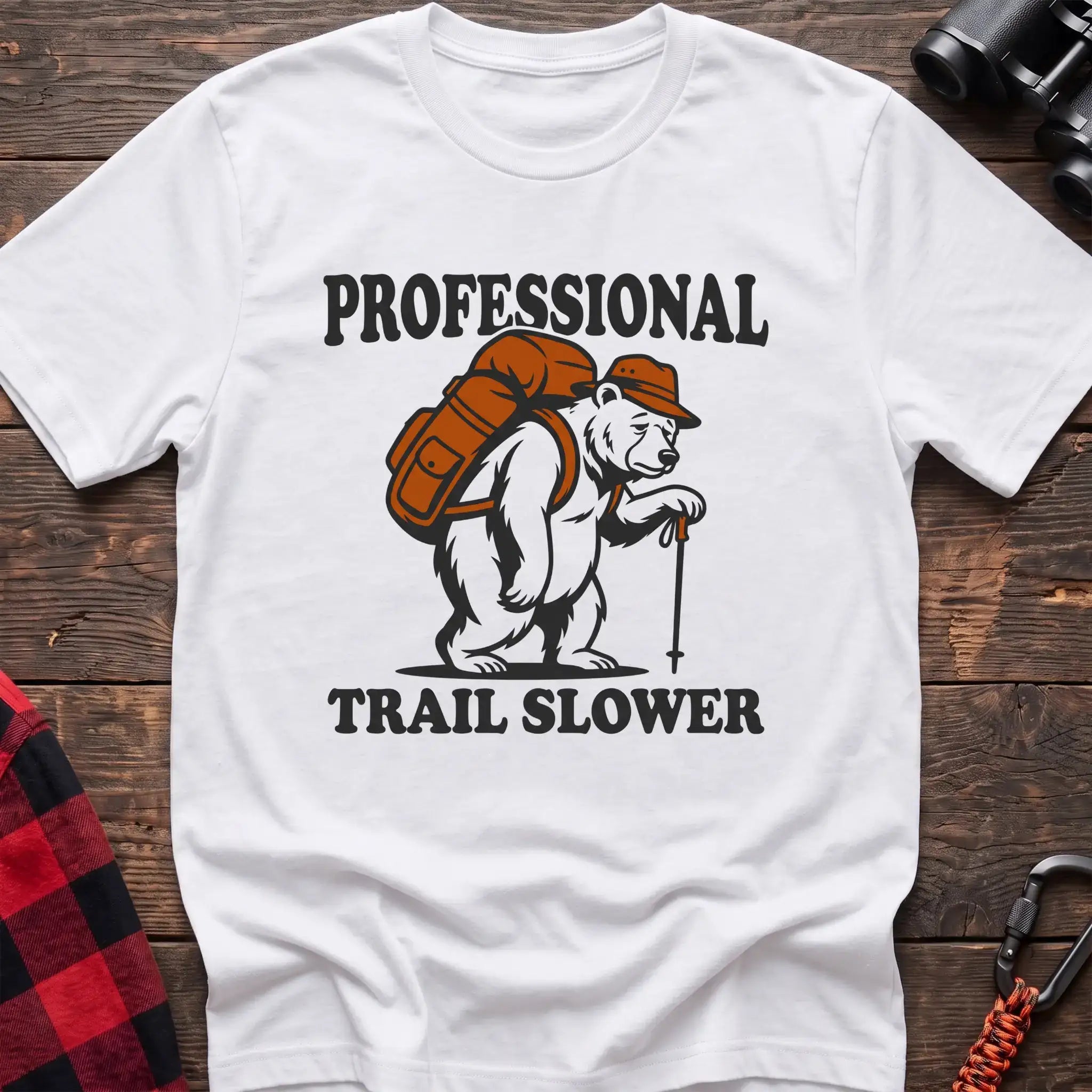 White t-shirt with a cartoon polar bear wearing a backpack and hat while holding a hiking stick, with the text “PROFESSIONAL TRAIL SLOWER,” laid flat on a wooden surface.