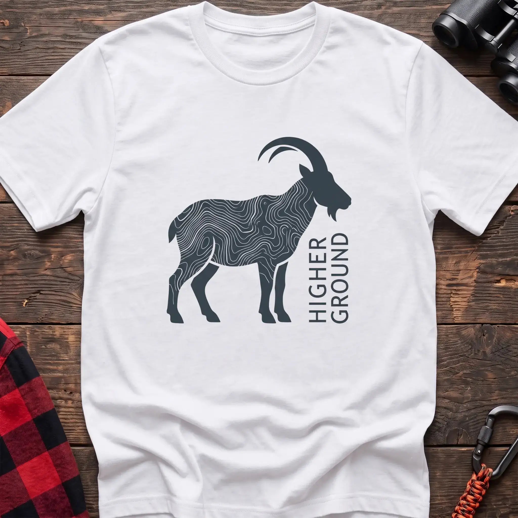 White t-shirt with a dark silhouette of a mountain goat filled with topographic contour lines, and the vertical text “HIGHER GROUND” printed beside it, laid flat on a wooden surface.
