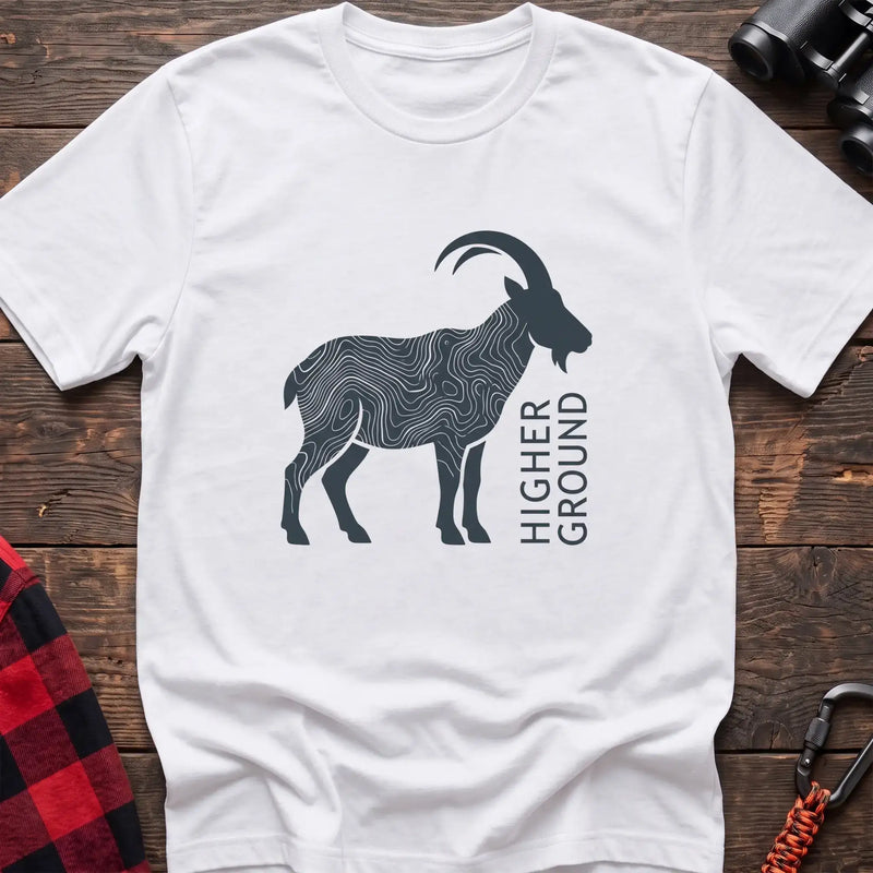 White t-shirt with a dark silhouette of a mountain goat filled with topographic contour lines, and the vertical text “HIGHER GROUND” printed beside it, laid flat on a wooden surface.