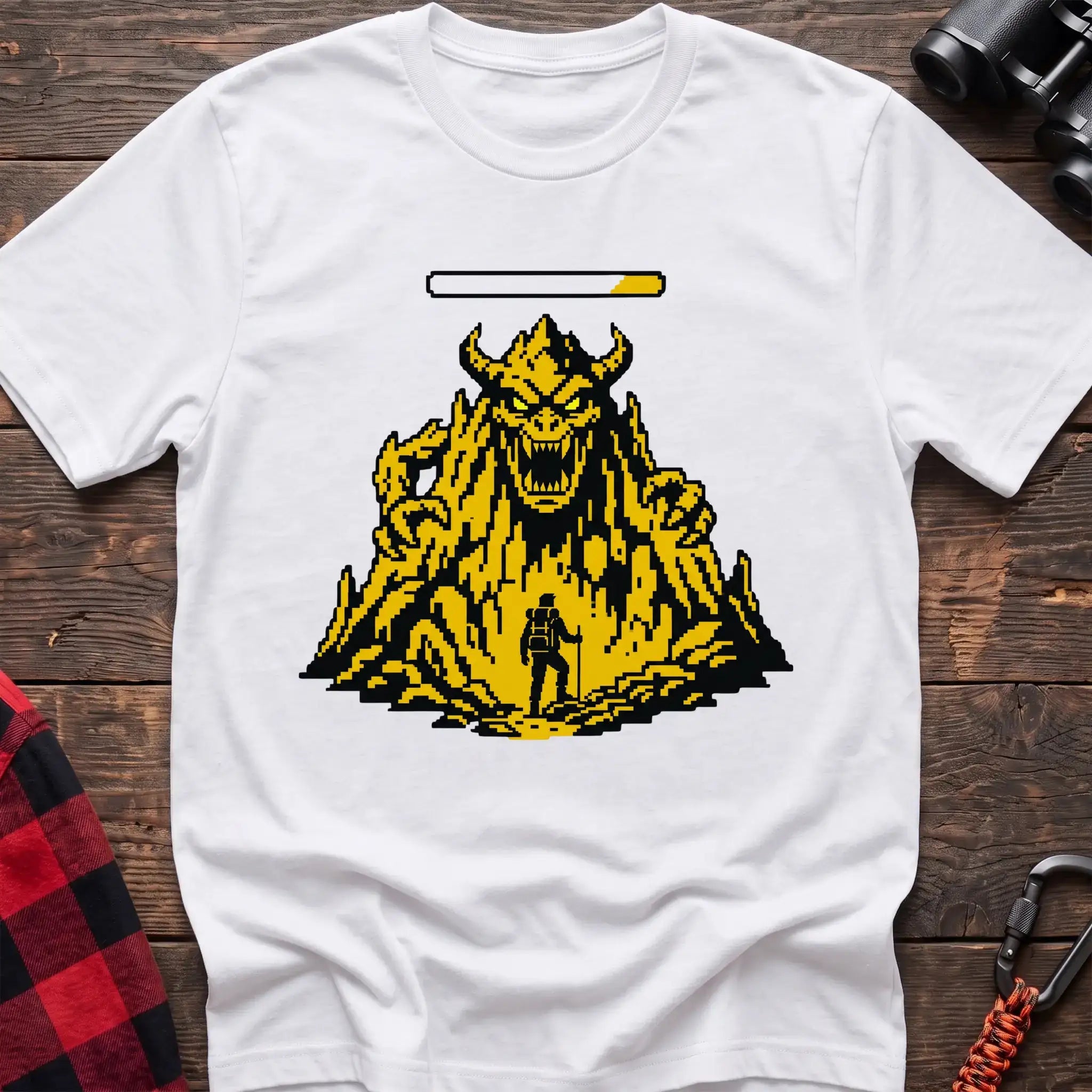 White t-shirt with a pixel-art illustration of a hiker facing a large horned mountain monster with a health bar above it, laid flat on a wooden surface.