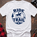 White cotton t-shirt with navy Ride the Trail graphic of a woman horseback riding through mountains and pine trees, flat lay on rustic wood with hiking gear