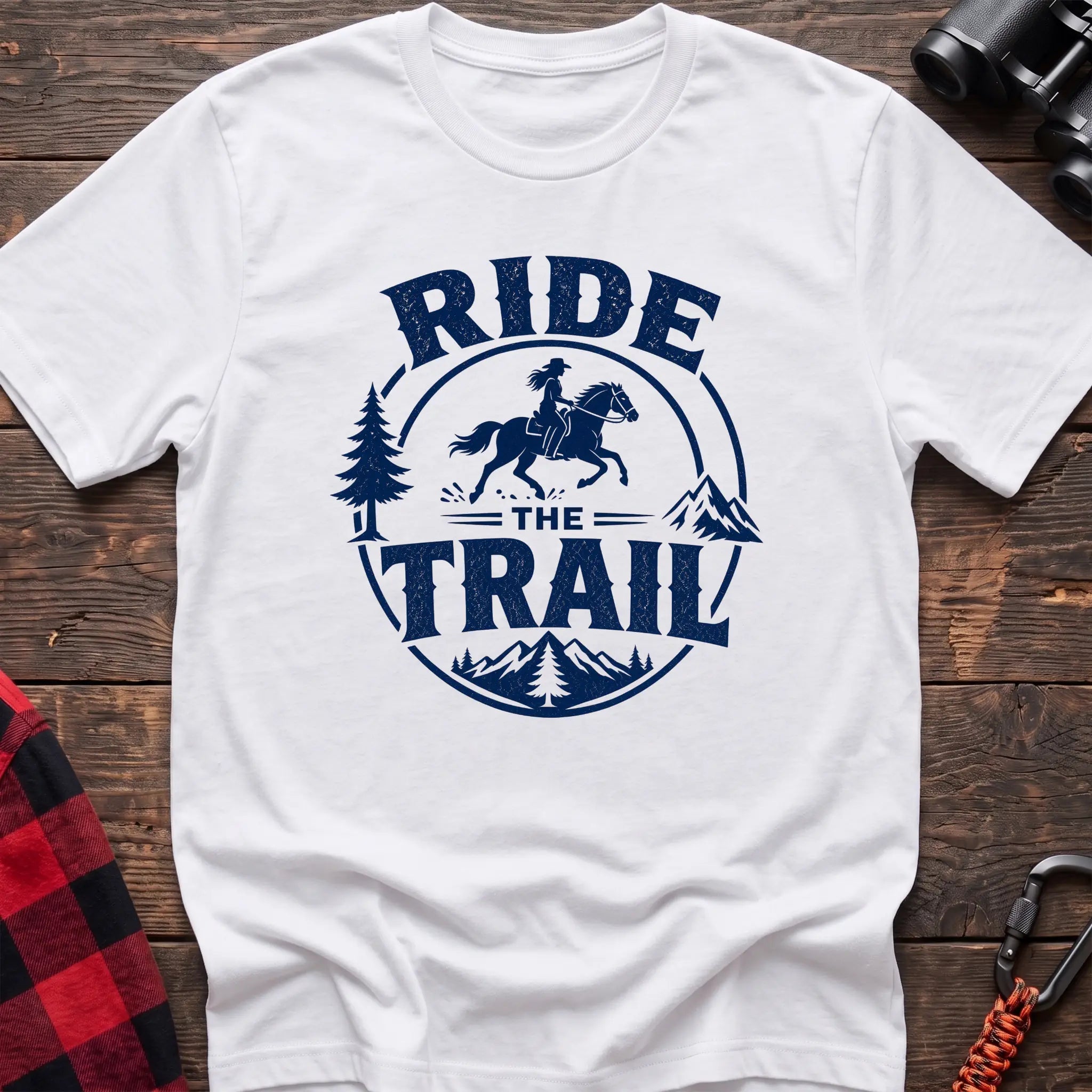 White cotton t-shirt with navy Ride the Trail graphic of a woman horseback riding through mountains and pine trees, flat lay on rustic wood with hiking gear
