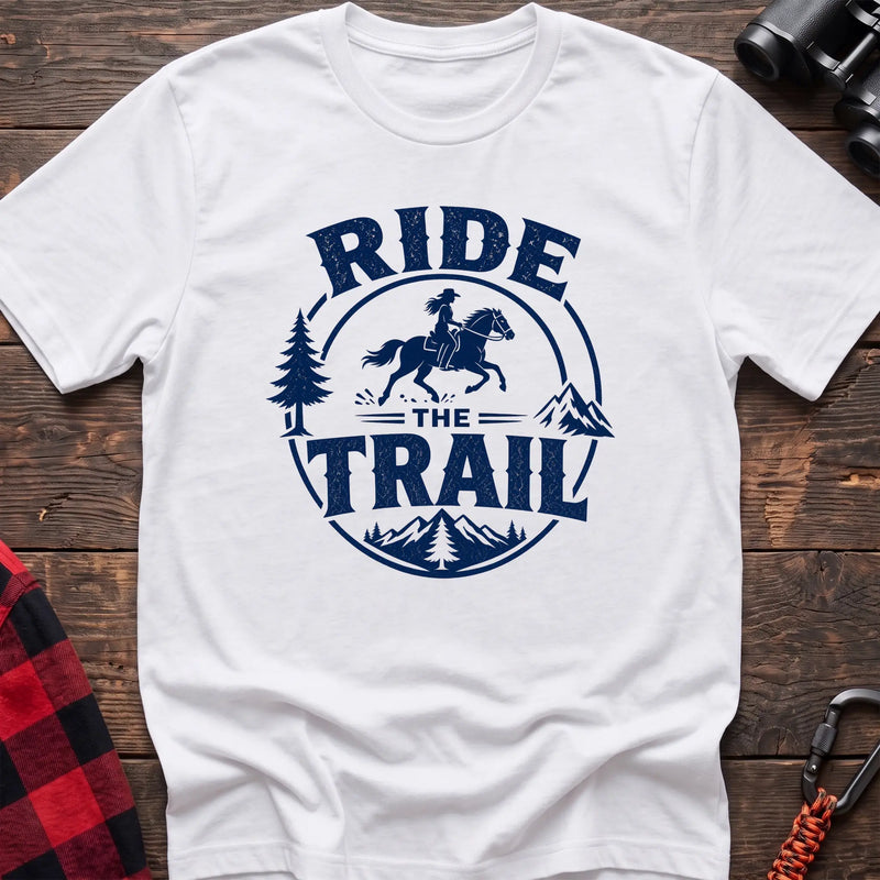 White cotton t-shirt with navy Ride the Trail graphic of a woman horseback riding through mountains and pine trees, flat lay on rustic wood with hiking gear