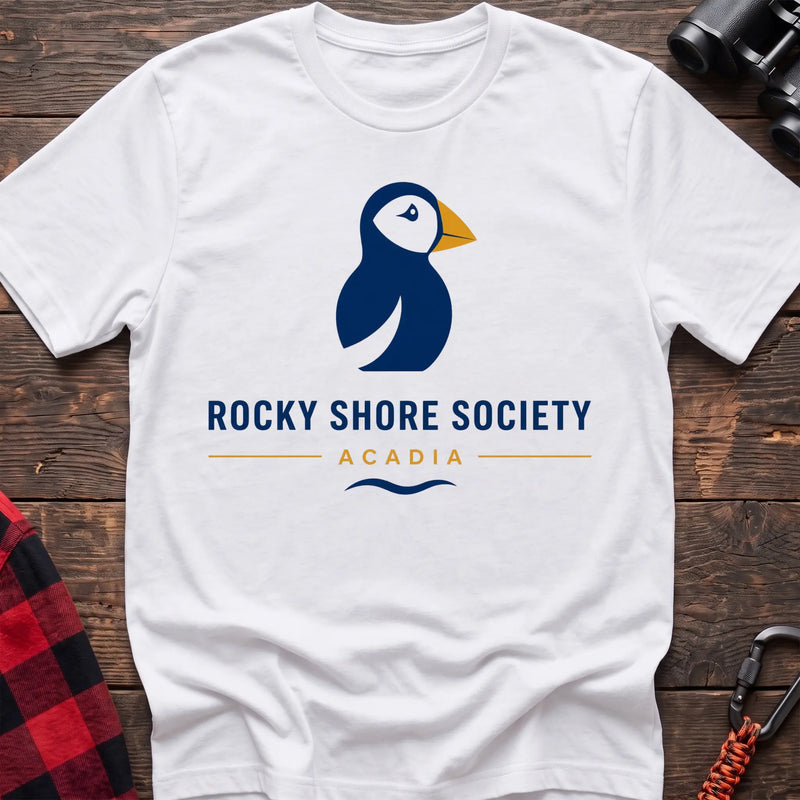 White cotton t-shirt with a navy puffin logo, yellow beak, and Rocky Shore Society Acadia text on a rustic wood background
