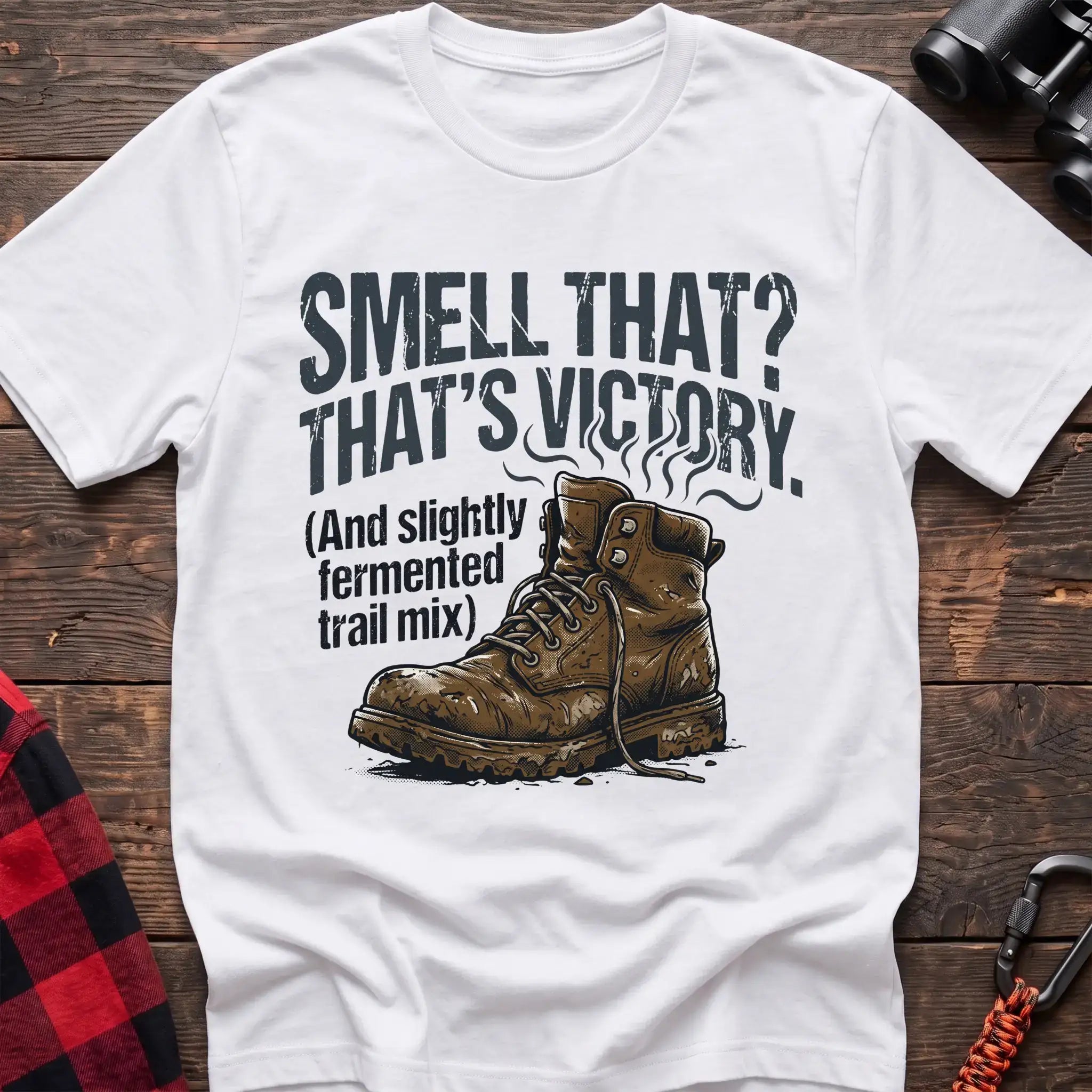 White t-shirt with an illustration of a dirty hiking boot with rising steam lines, and the text “SMELL THAT? THAT’S VICTORY. (And slightly fermented trail mix),” laid flat on a wooden surface