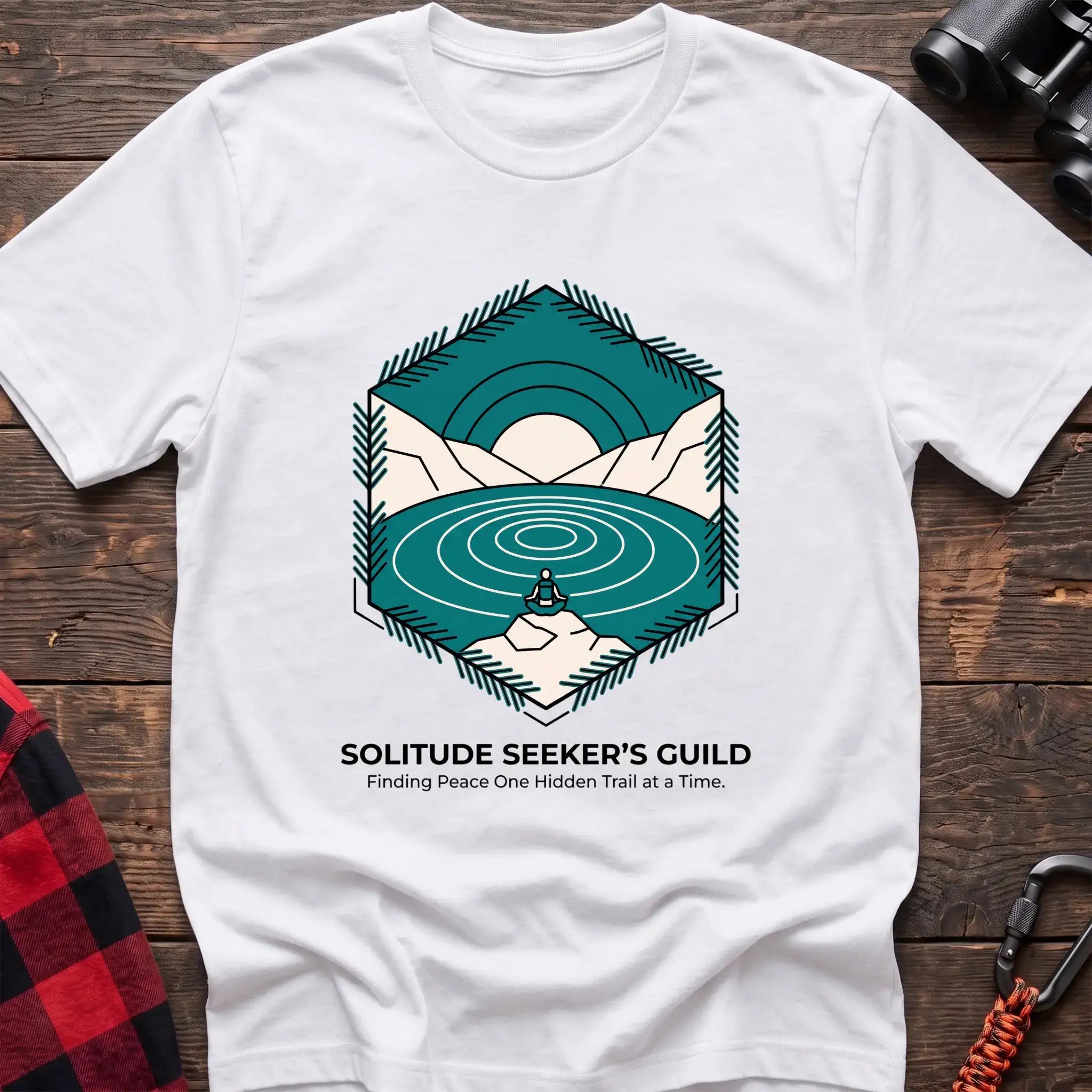 White t-shirt with a hexagonal illustration of a person meditating by a mountain lake, with the text “SOLITUDE SEEKER’S GUILD” and “Finding Peace One Hidden Trail at a Time,” laid flat on a wooden surface.