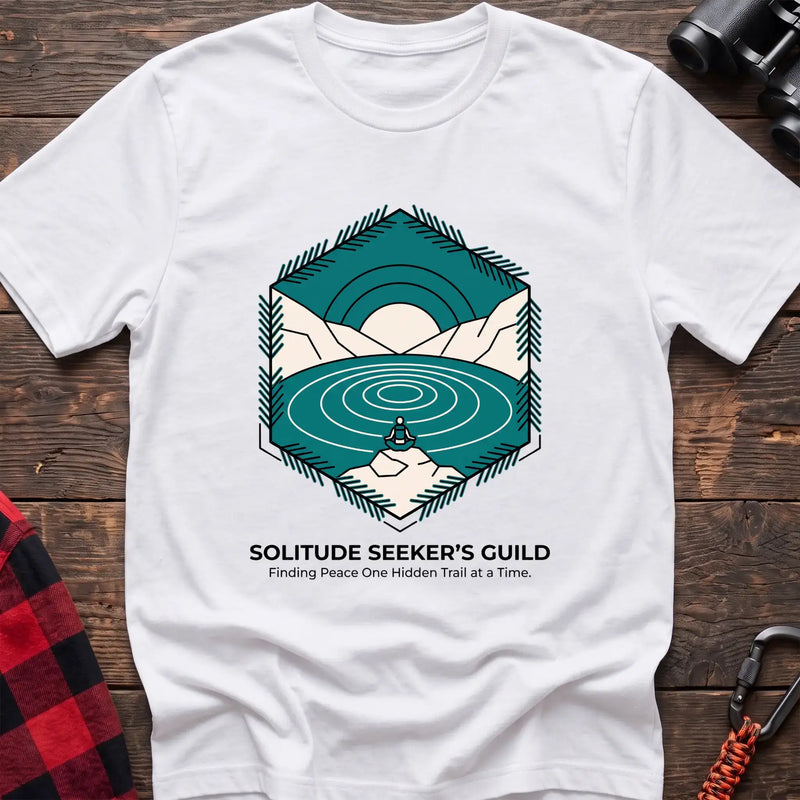 White t-shirt with a hexagonal illustration of a person meditating by a mountain lake, with the text “SOLITUDE SEEKER’S GUILD” and “Finding Peace One Hidden Trail at a Time,” laid flat on a wooden surface.