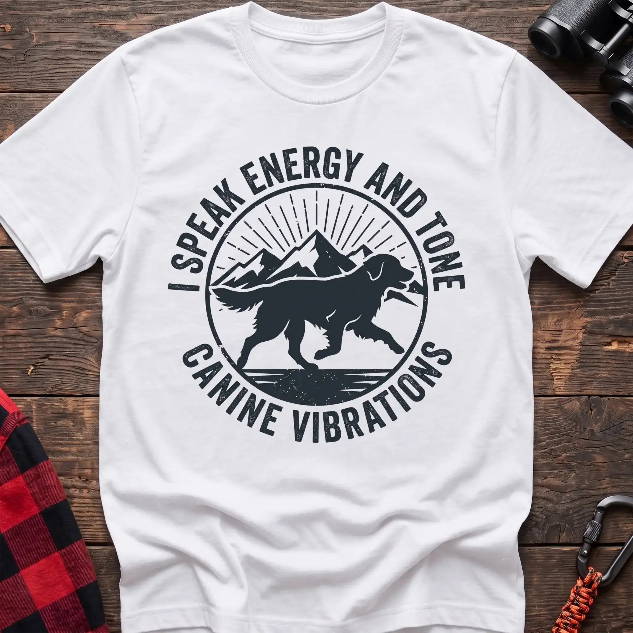 White t-shirt with a circular dark gray badge design featuring a running dog carrying a stick against mountain peaks and sun rays, with the text “I SPEAK ENERGY AND TONE” and “CANINE VIBRATIONS,” laid flat on a wooden surface.