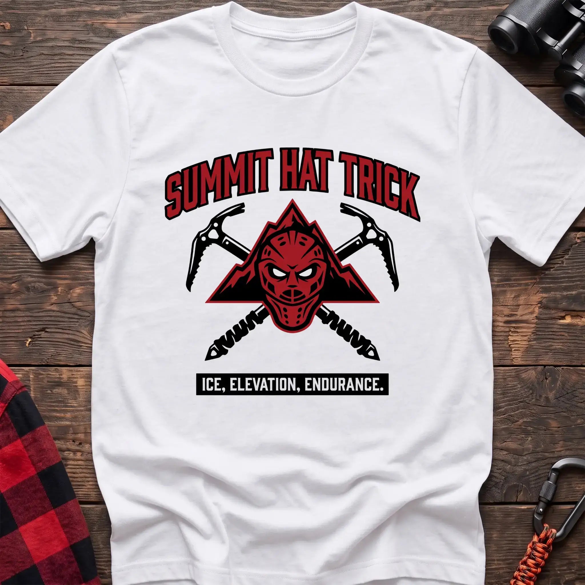 White t-shirt with a red mountain-themed goalie mask and crossed ice axes, with the text “SUMMIT HAT TRICK” and “ICE, ELEVATION, ENDURANCE,” laid flat on a wooden surface.