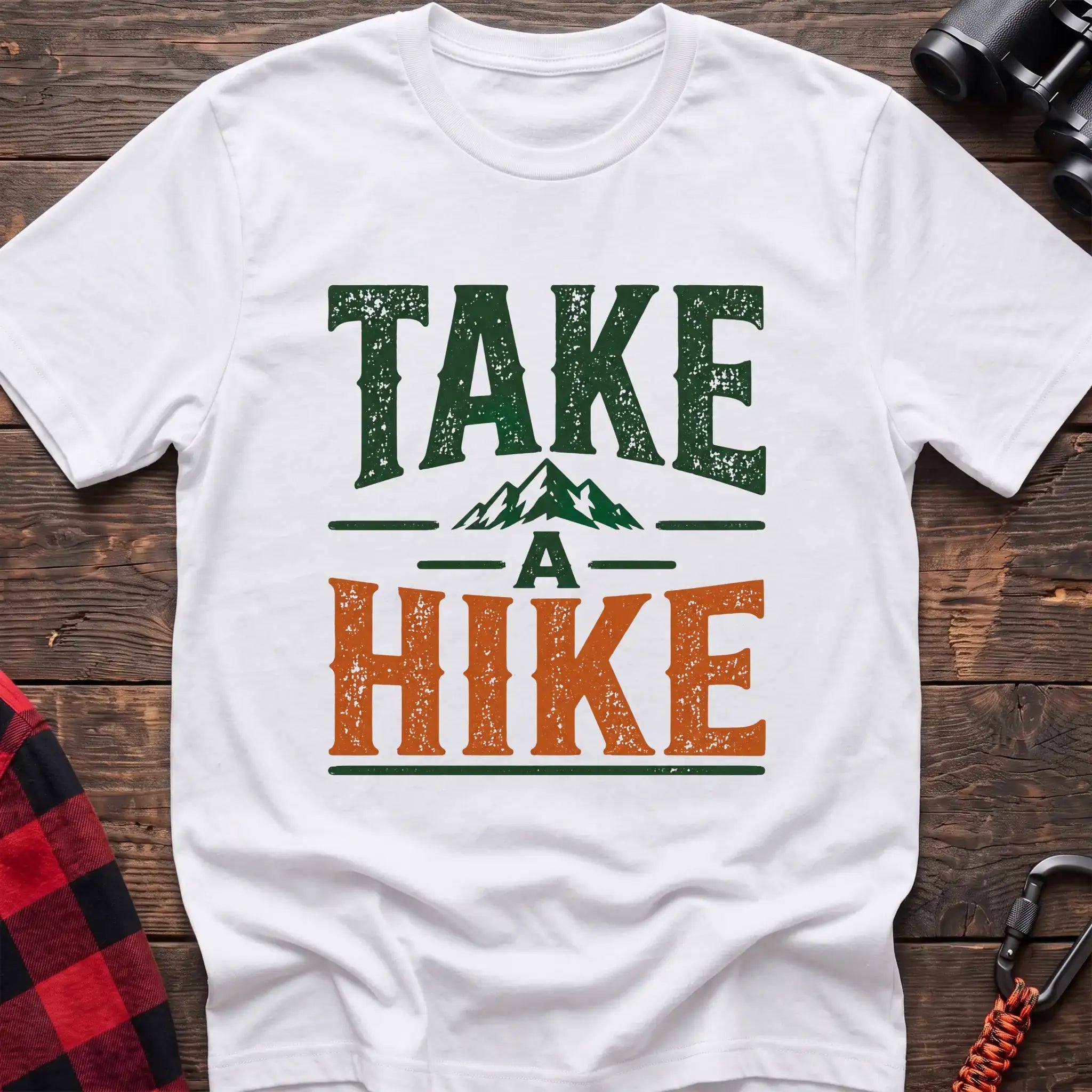 White t-shirt with distressed green and orange text reading “TAKE A HIKE” with a small mountain graphic between the words, laid flat on a wooden surface.