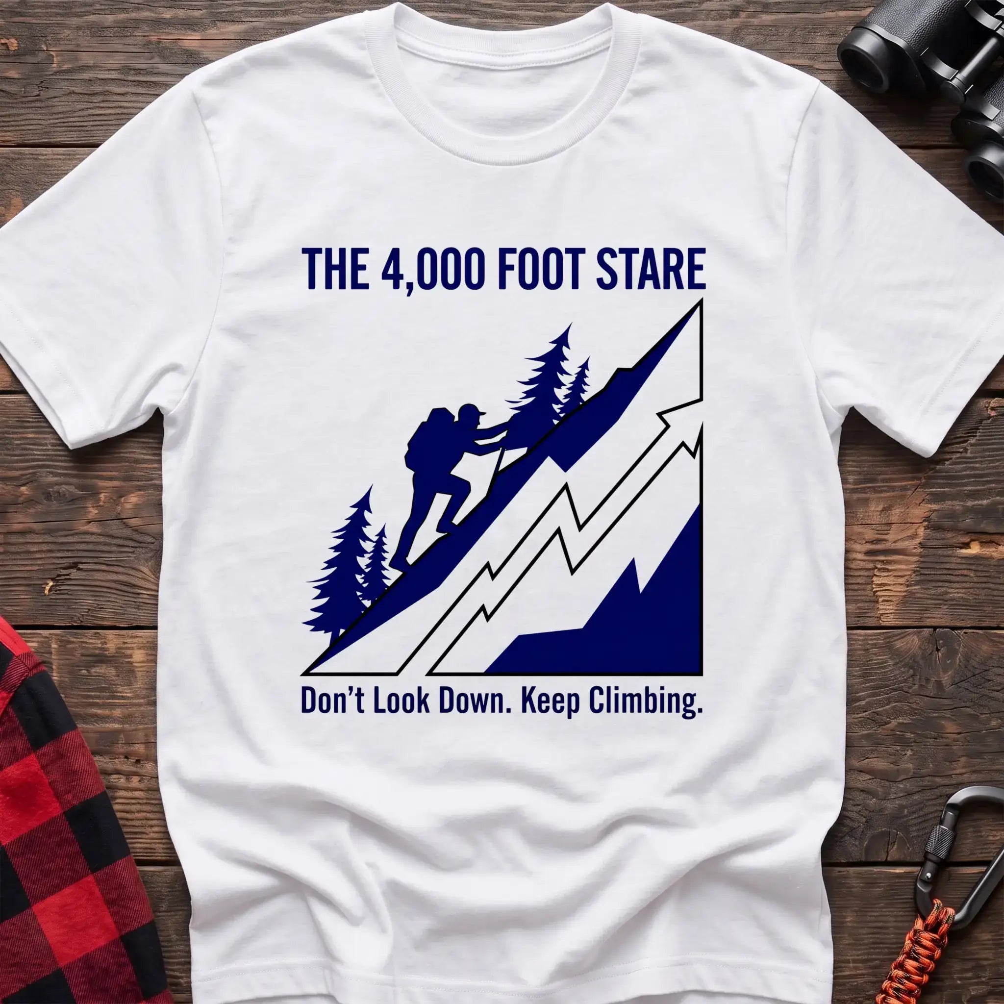 White t-shirt with a blue illustration of a hiker climbing a steep mountain, with the text “THE 4,000 FOOT STARE” and “Don’t Look Down. Keep Climbing,” laid flat on a wooden surface.
