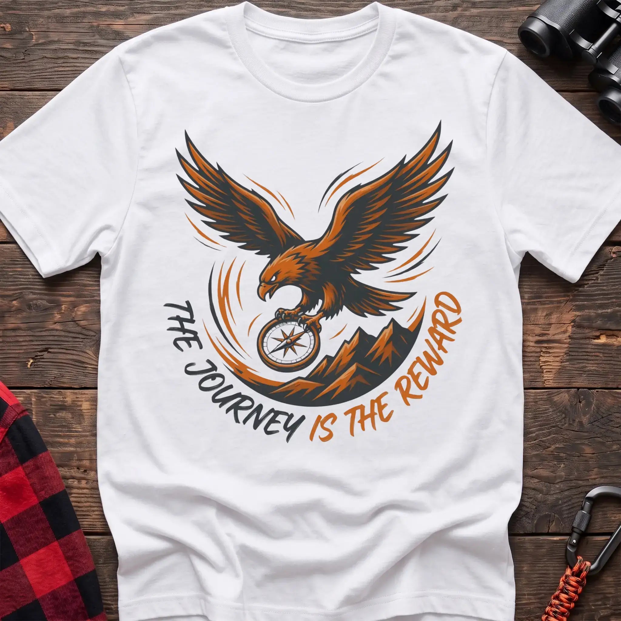 White t-shirt with an orange and dark graphic of a soaring eagle holding a compass above mountains, with the curved text “THE JOURNEY IS THE REWARD,” laid flat on a wooden surface.