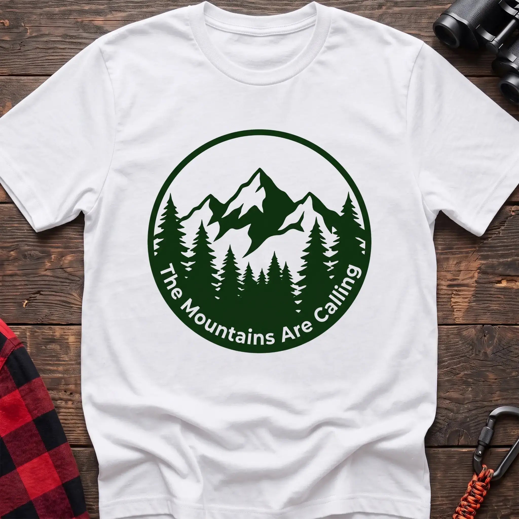 White t-shirt with a circular dark green design featuring mountain peaks and pine trees, with the text “The Mountains Are Calling,” laid flat on a wooden surface.