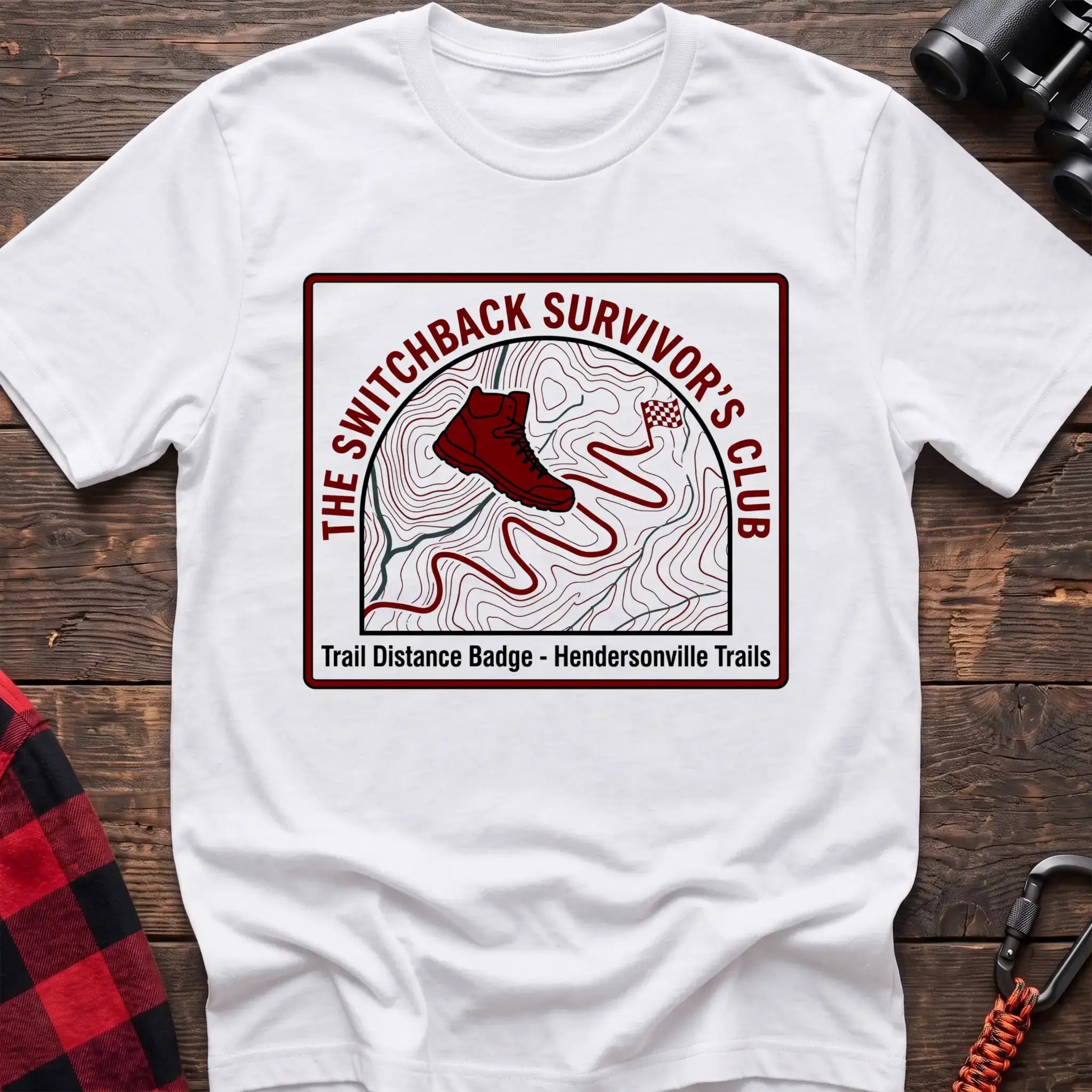 White t-shirt with a square badge design featuring a red hiking boot on a topographic map with a winding trail, and the text “THE SWITCHBACK SURVIVOR’S CLUB” and “Trail Distance Badge - Hendersonville Trails,” laid flat on a wooden surface.