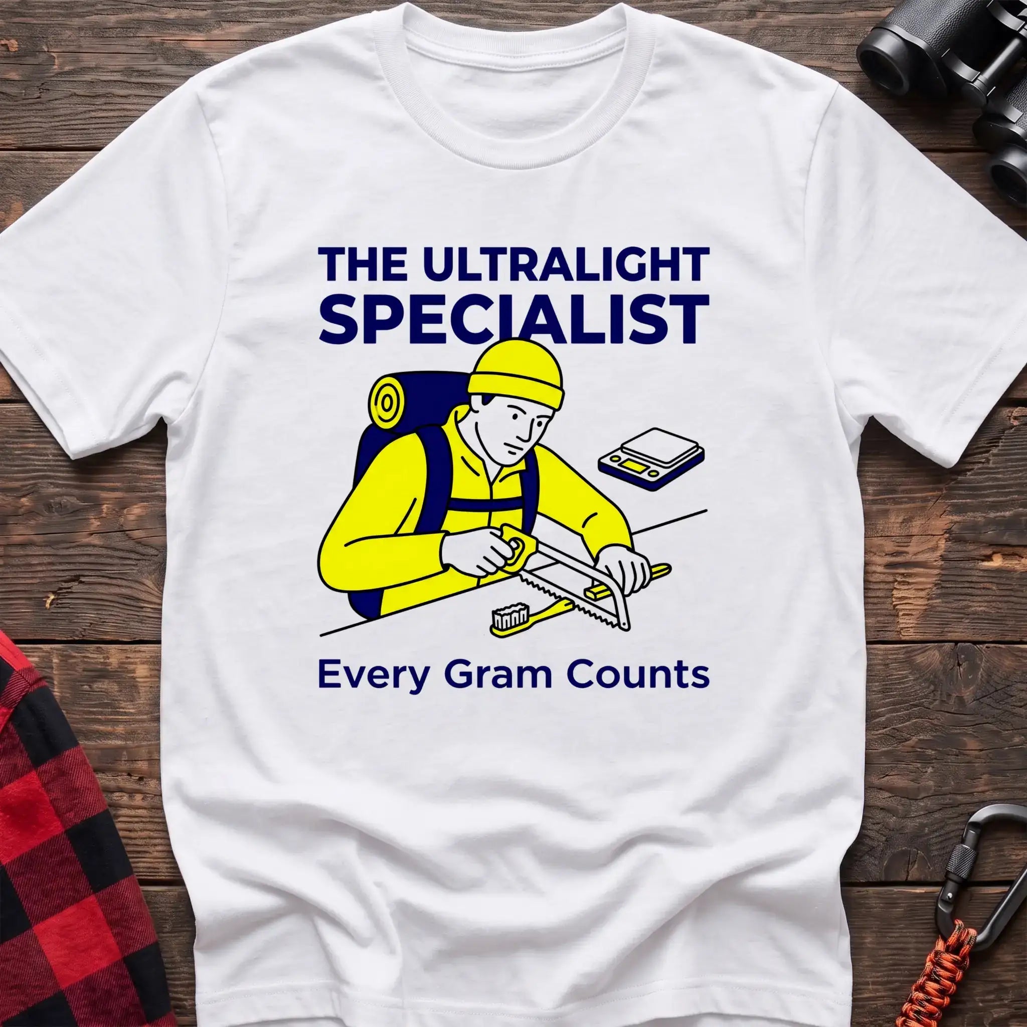 White t-shirt with an illustration of a hiker sawing a toothbrush on a table next to a scale, with the text “THE ULTRALIGHT SPECIALIST” and “Every Gram Counts,” laid flat on a wooden surface.