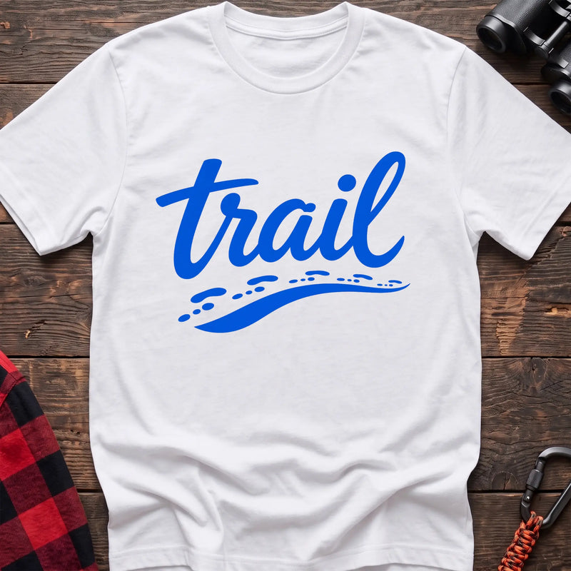 White cotton t-shirt with blue cursive trail script and dotted path graphic, flat lay on rustic wood with hiking gear