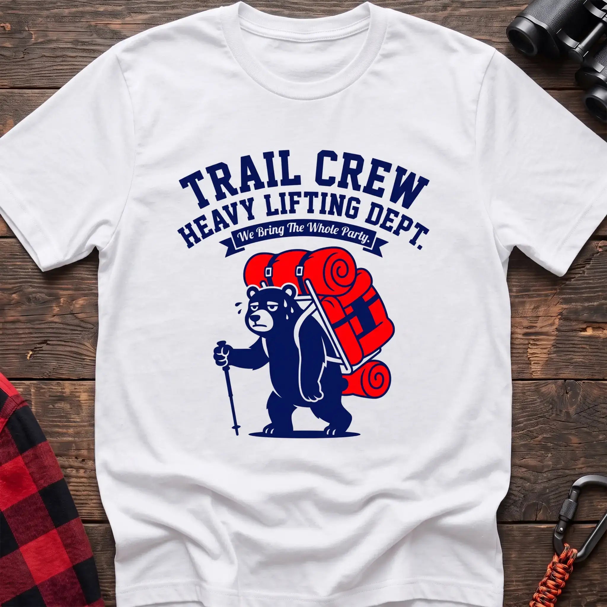 White t-shirt with a cartoon dark blue bear carrying a large red backpack and hiking pole, with the text “TRAIL CREW HEAVY LIFTING DEPT. We Bring The Whole Party,” laid flat on a wooden surface.