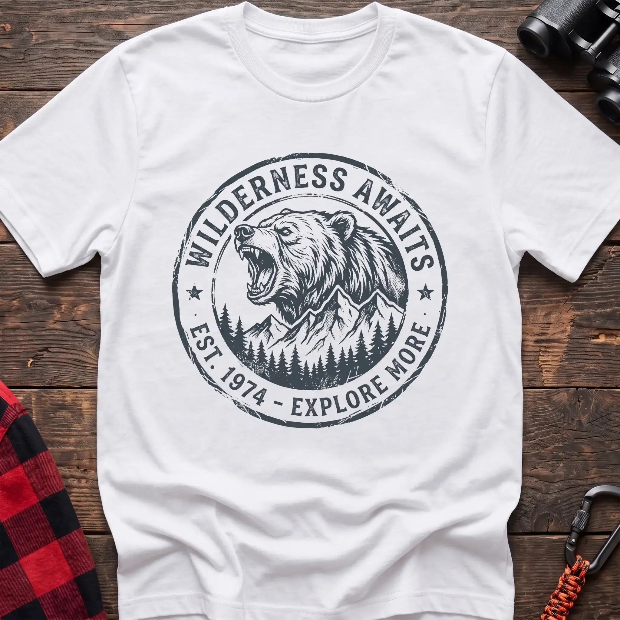 White t-shirt with a circular dark gray badge design featuring a roaring bear head above mountains and pine trees, with the text “WILDERNESS AWAITS” and “EST. 1974 - EXPLORE MORE,” laid flat on a wooden surface.