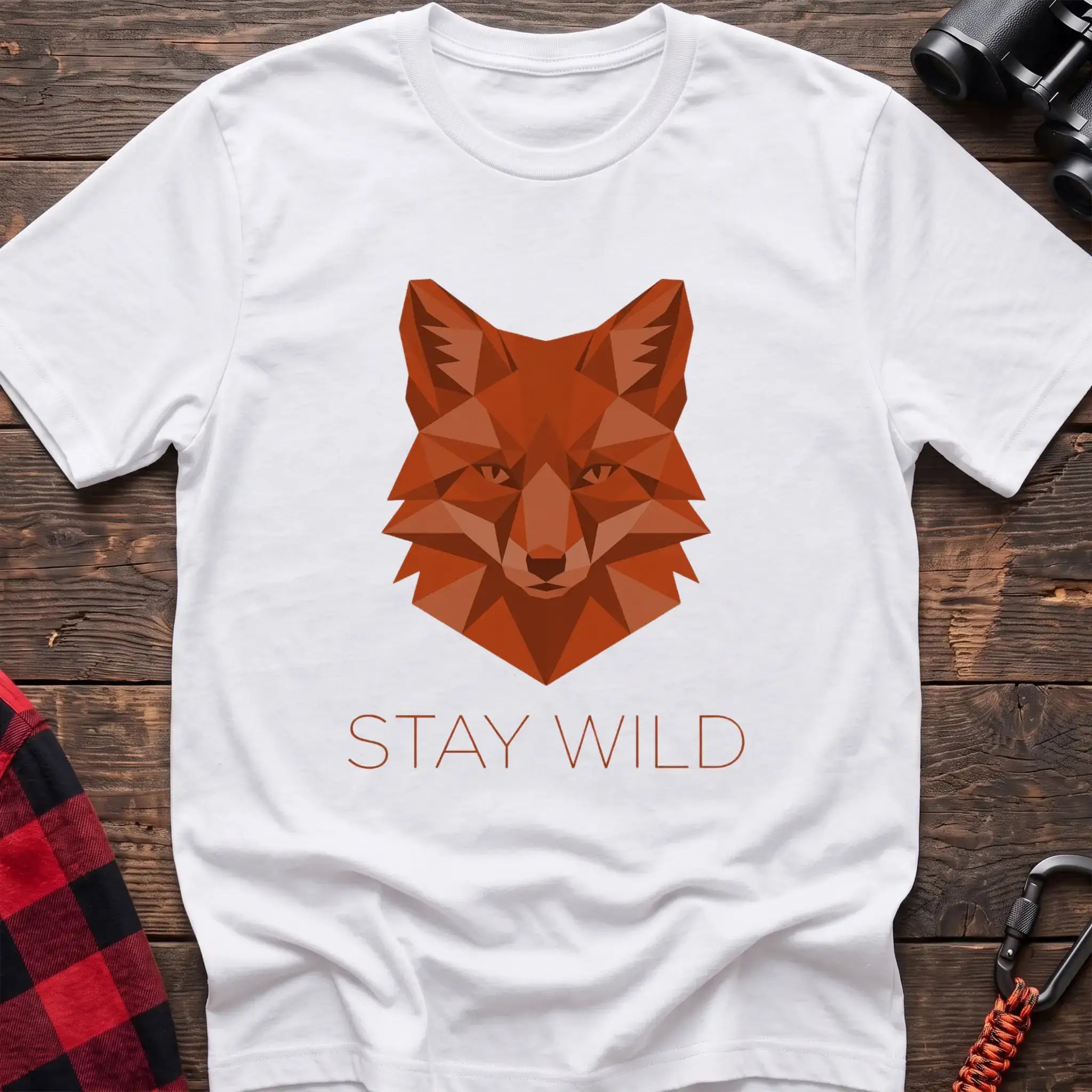 White t-shirt with a geometric low-poly orange fox head and the text “STAY WILD” printed below in matching orange, laid flat on a wooden surface.
